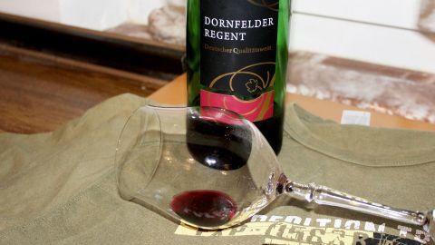 Ein T-Shirt liegt auf einem Tisch, während ein Glas mit Rotwein daneben steht, was einen möglichen Rotweinfleck zeigt.