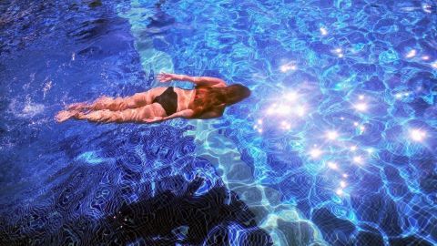 Eine Frau schwimmt entspannt im blauen Wasser eines Schwimmbades, umgeben von glitzernden Lichtreflexen.