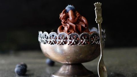 Eine silberne Schale enthält samtige Mousse au Chocolat, dekoriert mit einer Beere, umgeben von blauen Beeren und löffelbereit.