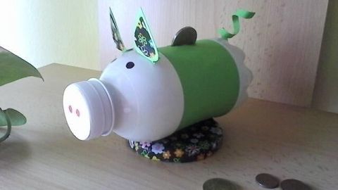 Ein DIY-Sparschwein aus einer PET-Flasche liegt dekoriert auf einem Tisch, umgeben von Münzen und einem Pflanzentopf.