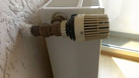 Ein Heizkörper-Thermostatventil ragt aus der Wand und ist an einen Heizkörper angeschlossen; Sonnenlicht fällt durch ein Fenster.