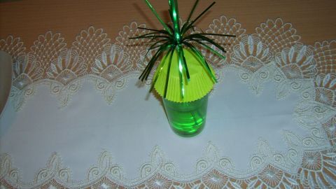 Ein grüner Becher mit gelber Abdeckung und einer Spinne aus Plastik schützt Getränke vor Insekten, auf einem weißen Tischläufer.