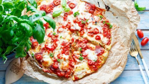 Käse und Tomatensauce bedecken eine Low Carb Pizza, die auf Backpapier liegt, umgeben von frischem Basilikum und einer Holzlöffel.