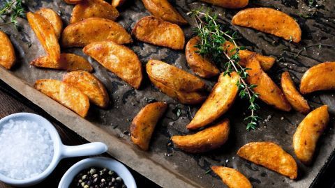 Knusprige Kartoffelspalten liegen auf einem Backblech, umgeben von frischen Kräutern und Schalen mit Salz und Pfeffer.