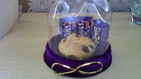Plätzchen unter PET-Flasche, umgeben von zwei Snack-Packungen, dekoriert mit violettem Stoff und goldener Schleife auf Tisch.