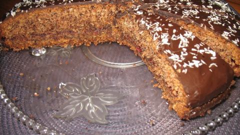 Schoko-Nuss-Torte mit Beerenfüllung liegt auf Glasplatte, ein Stück fehlt, Schokolade und Kokosraspeln dekorieren die Oberfläche.
