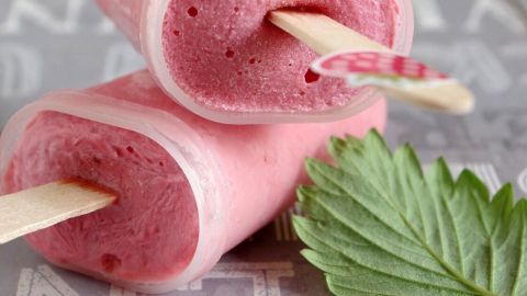 Die rosa Joghurt-Früchte-Eis am Stiel liegt auf einem grau gemusterten Untergrund, umgeben von einem frischen Blatt.