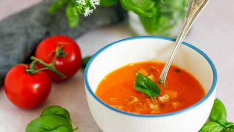 Ein Teller mit kalter Tomatensuppe wird von einem Löffel umgerührt, umgeben von frischen Tomaten und Kräutern.