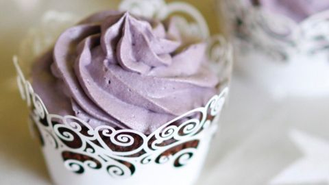 Kleine, dekorative Muffins mit lila Sahnehaube werden auf einer hellen, gemusterten Tischdecke präsentiert.