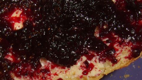 Eine dunkle, glänzende Schwarze Johannisbeer-Konfitüre breitet sich über ein frisch gebackenes Brot aus, das auf einem blauen Teller liegt.