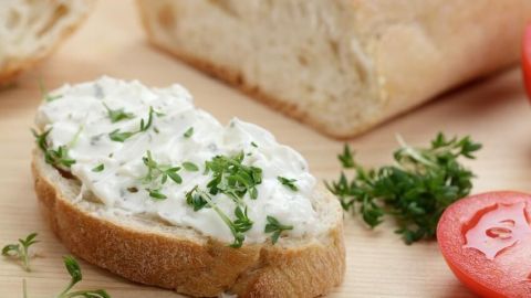 Eine Scheibe Brot ist mit Frischkäse und Kresse bestrichen; im Hintergrund liegt Brot, daneben eine Tomate.
