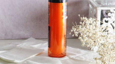 Ein Flakon mit orangefarbener Flüssigkeit steht auf weißen Tüchern, umgeben von einer blassen Hintergrunddekoration.