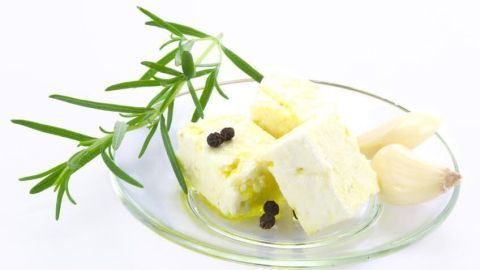 Frische Käsewürfel liegen auf einem Glasgericht, umgeben von Kräutern und Knoblauchzehen, bereit zur Verwendung.