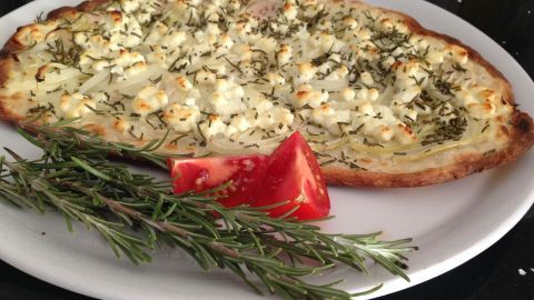 Ein vegetarischer Flammkuchen liegt auf einem weißen Teller, dekoriert mit Ziegenkäse und frischen Kräutern.