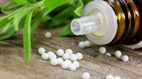 Kleine weiße Kügelchen liegen neben einer braunen Flasche mit homöopathischem Mittel und frischen Kräutern auf Holz.