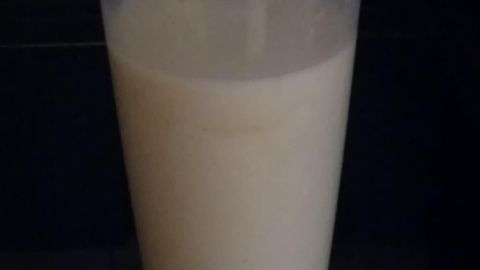 Ein Glas mit Bananensaft steht auf einem Tisch, umgeben von einem dunklen Hintergrund. Es wirkt frisch zubereitet.