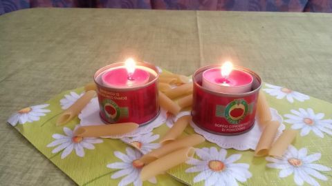 Zwei Kerzen in Dosen von Tomatenmark brennen, umgeben von gelben Blüten und auf einer fröhlichen Tischdecke im Hintergrund.