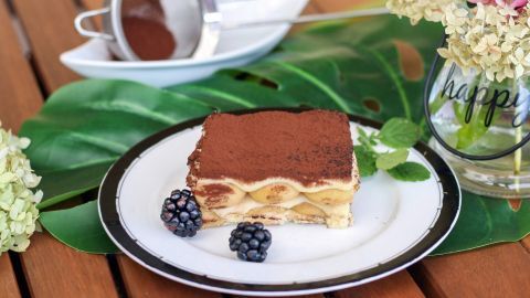 Tiramisu ohne Alkohol serviert auf einem Teller mit Brombeeren neben einem Sieb auf Holztisch, umgeben von Blumen und Blättern.