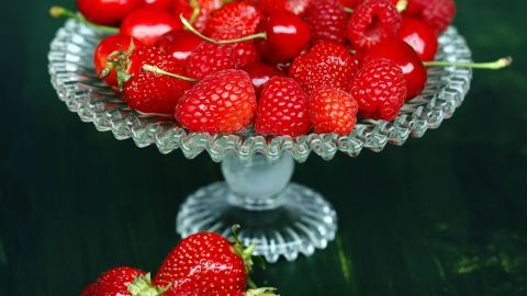 Ein dekorativer Glasschalen voller frischer Erdbeeren steht auf einem grünen Tisch, umgeben von weiteren Erdbeeren.
