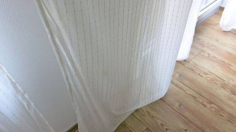 Leichte, weiße Gardinen hängen vor einem Fenster und bewegen sich sanft im Licht eines hellen Raumes mit Holzfußboden.
