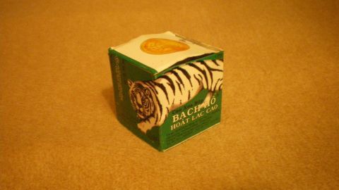 Ein kleiner, quadratischer Tigerbalsam-Behälter steht auf einem flachen, braunen Untergrund und ist für die Behandlung von Warzen gedacht.