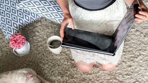 Eine Person hält ein Fotobuch in den Händen, während sie auf einem Teppich sitzt. Neben ihr liegt ein Hund und eine Tasse steht bereit.