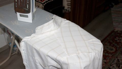 Ein Hemd liegt auf einem Bügelbrett, während ein Bügeleisen bereit zum Bügeln daneben steht, in einem wohnlichen Raum.