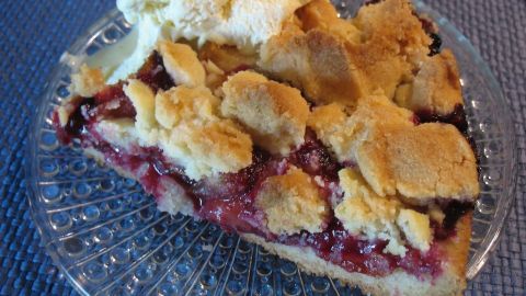 Eine köstliche Zwetschgen-Streusel-Torte auf einem Glasteller, serviert mit einer Portion Sahne, auf einem blauen Tisch.