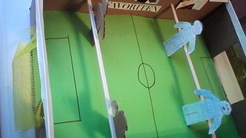 Ein Tischkicker aus Karton mit grünem Spielfeld und Figuren in Aktion, platziert auf einem Tisch in einem kreativen Raum.