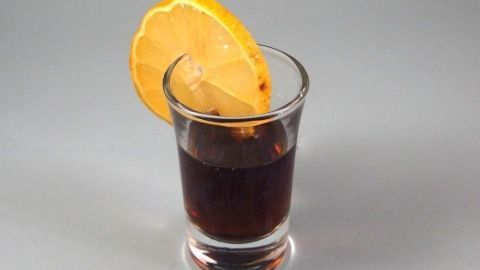Ein kleiner, dunkler Getränkeglas mit Ramazzotti, garniert mit einer frischen Zitronenscheibe, steht auf einem grauen Untergrund.