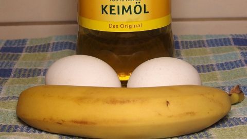 Eine Banane liegt auf einem karierten Tuch, während zwei Eier und eine Flasche Bananen-Öl im Hintergrund stehen.