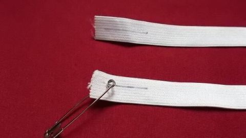 Ein Gummiband wird mit einer Sicherheitsnadel in einen Bund eingeführt, auf einem roten Untergrund.