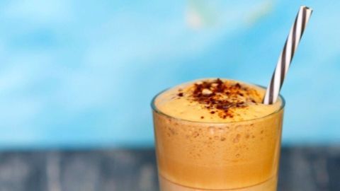 Ein Glas Cappuccino-Eiskaffee mit Schaumkrone und Kakaopulver wird in einer lichtdurchfluteten Umgebung präsentiert.