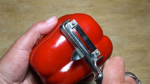 Eine Hand schält eine rote Paprika mit einem Schäler auf einer Holzoberfläche, um sie bekömmlicher zu machen.