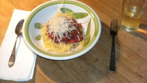 Spaghetti mit Tomatensoße und geriebenem Käse in einer Schale auf Holztisch, daneben ein Glas Getränk und Besteck.