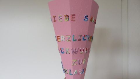 Eine pinke Mini-Schultüte wird mit bunten Buchstaben verziert, während sie in einer Hand gehalten wird, vor einer Wand.