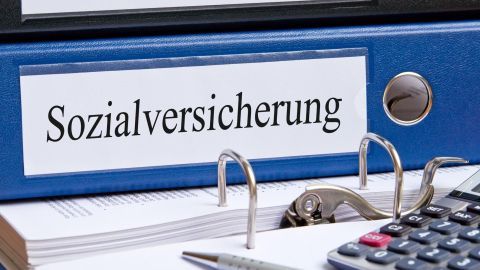 Ein blauer Ordner mit der Aufschrift „Sozialversicherung“ liegt auf einem Tisch neben einem Taschenrechner.