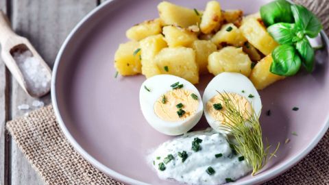 Auf einem Teller liegen gekochte Eier und grüne Soße neben knusprigen Kartoffeln, umgeben von einem naturbelassenen Untergrund.