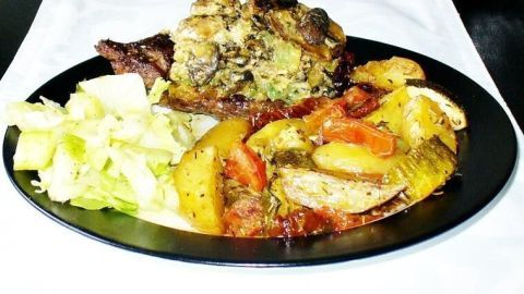 Überbackene Steaks mit einer Käse-Pilz-Haube liegen auf einem schwarzen Teller, umgeben von buntem Ofengemüse und Salat.
