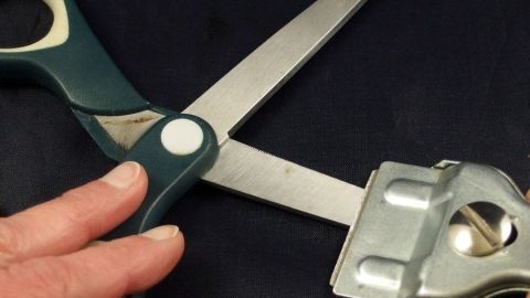 Eine Schere wird mit einem Werkzeug in der Hand gehalten, um Klebereste zu entfernen, vor einem dunklen Hintergrund.