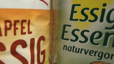 Zwei Flaschen Essig stehen nebeneinander, eine für Apfelessig, die andere für Essigessenz, beide in heller Umgebung.