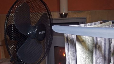 Ein Wäscheständer mit feuchter Wäsche steht neben einem Ventilator, der eine warme, kleine Wohnumgebung unterstützt.
