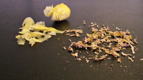 Hauchdünn geschälte und geschnittene Ingwerstücke liegen auf einer dunklen Oberfläche neben Schalenresten.