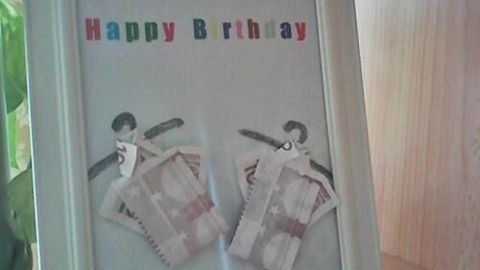 Bilderrahmen zeigt gefaltete Geldscheine als Geschenk, dekoriert mit dem Text "Happy Birthday", auf einem Holzregal.