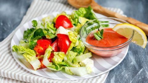 Ein frischer Salat mit Tomaten und grünen Blättern liegt auf einem Teller, daneben ein Glas mit amerikanischem Salatdressing und einer Zitrone.