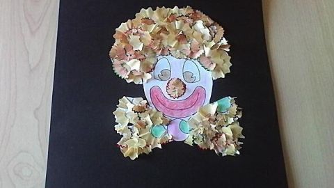 Ein Clown aus Buntstiftspänen ziert eine schwarze Pappe im Kinderzimmer, mit buntem Gesicht und lockigem Haar.