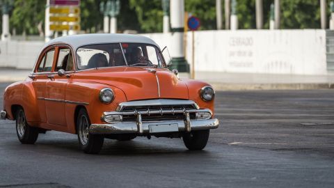 Ein orangefarbenes Auto fährt langsam auf einer Stadtstraße, während Vogelkot am Autolack sichtbar eingebrannt ist.