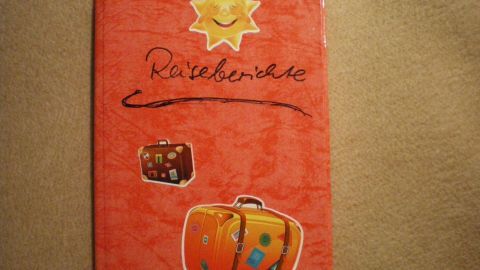Ein rotes Notizbuch mit der Aufschrift „Reiseberichte“ liegt auf einem neutralen Untergrund und zeigt bunte Reisegepäck-Aufkleber.