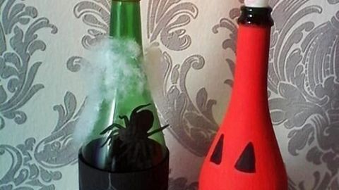 Zwei dekorierte Flaschen stehen auf einem Tisch; eine rote Flasche mit einem grinsenden Gesicht und eine grüne mit Kerze.