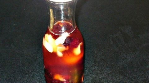 Eine Glasflasche mit fruchtigem Brombeeressig und Ingwer steht auf einem schwarzen Tisch, umgeben von getrockneten Brombeeren und Nüssen.